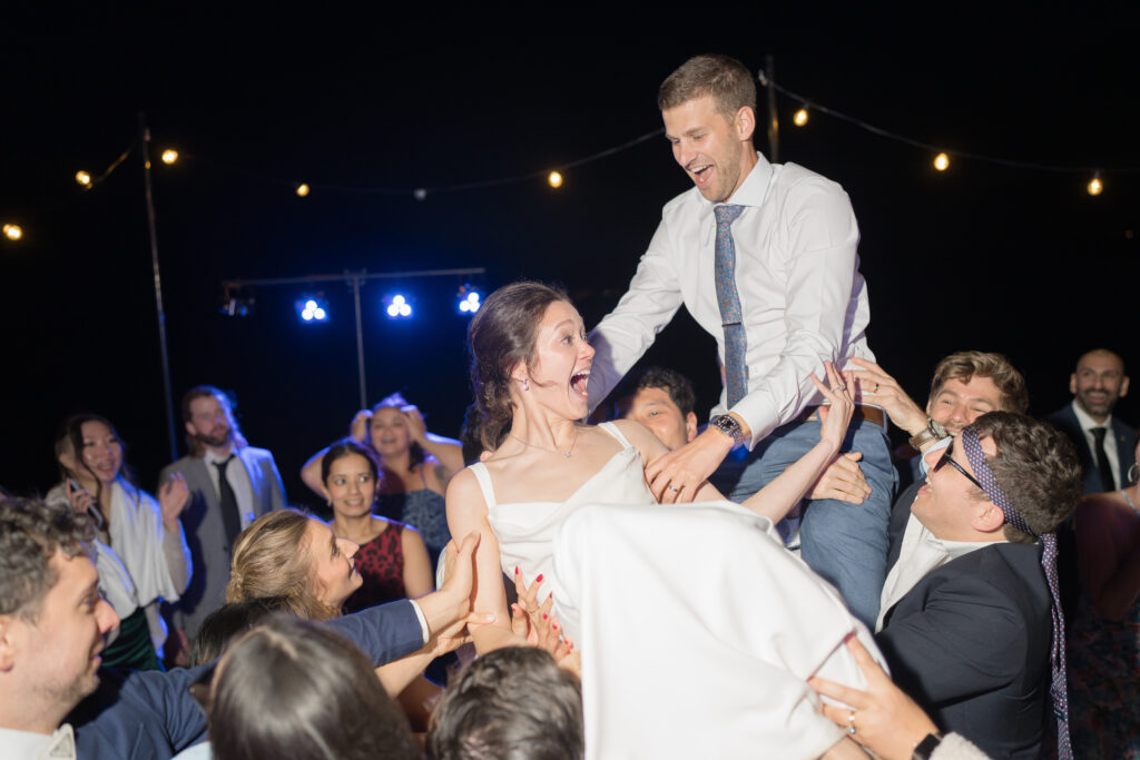 The bride and groom are lifted into the air by friends during a joyful dance party at their Hope Island Beach Club wedding in La Conner, planned by Prologue Events. Guests cheer around them under glowing string lights.